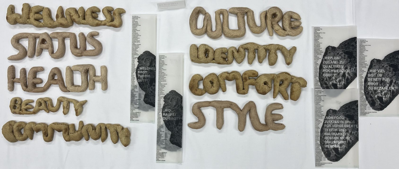 installation: baked bread letters spelling wellness, status, health, beauty, community, culture, identity, comfort and style, laid out on a white table between the printed bread bags