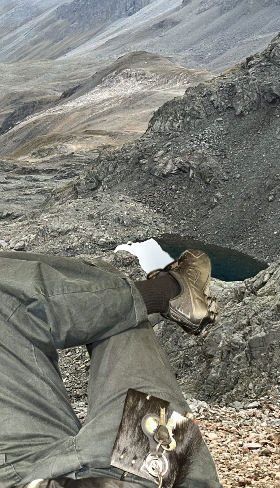 advertising image: legs in grey trousers crossed on a rocky mountain slope above a small dark lake, a black cowhide charm with keys and carabiner hanging from the pocket