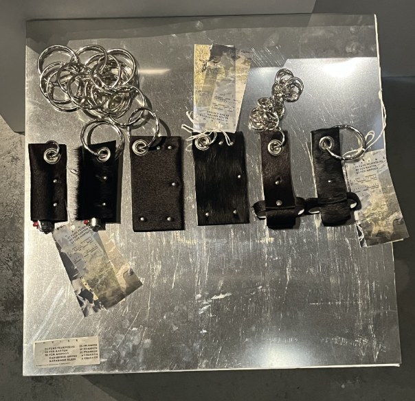 the finished charms in black cowhide with rivets, eyelets and carabiners, laid out on a steel plate with their tracing paper labels and the price list