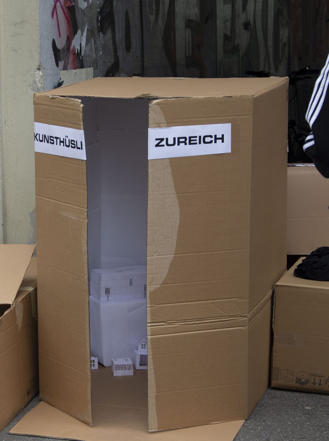 a large cardboard box standing on the street, labelled KUNSTHÜSLI and ZUREICH, opened to show a white interior with small paper buildings