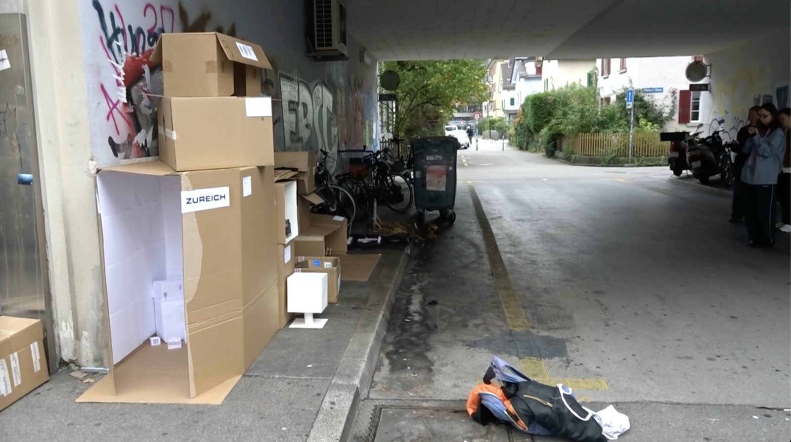 the Zureich box among stacked cardboard boxes in a graffiti covered underpass, next to bikes and a bin
